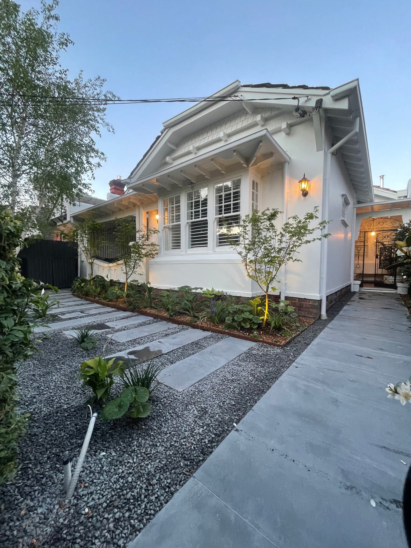 A three-week front garden transformation. Bluestone steppers lead to the entrance through silver gravel, with a planting palette built for texture and shade — leopard plants, silver lady ferns, a Japanese maple (Acer palmatum), Fatsia japonica, evergreen giant liriope, sea lavender for colour and NZ rock lilies for contrast. Finished with a custom black timber privacy screen and a monument grey bin holder.